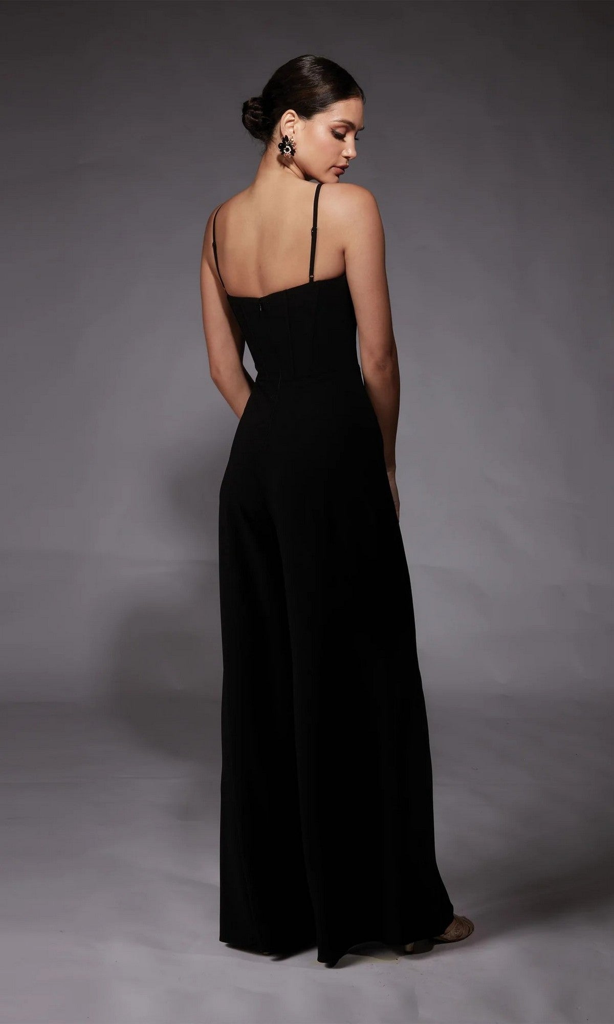 A woman in a black Long Formal Jumpsuit: Alyce 70075, featuring a sleeveless, corset-boned bodice, stands with her back to the camera against a gray background.